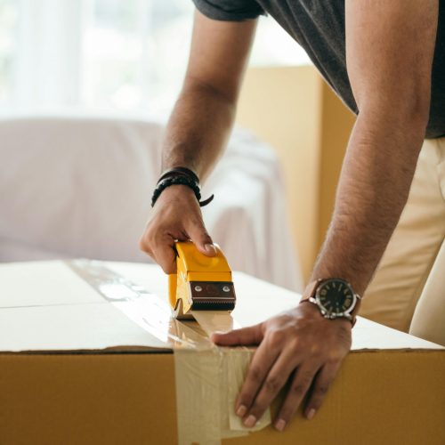 Side view of concentrated crop ethnic male sticking cardboard box with tape during preparing for moving in house at daytime